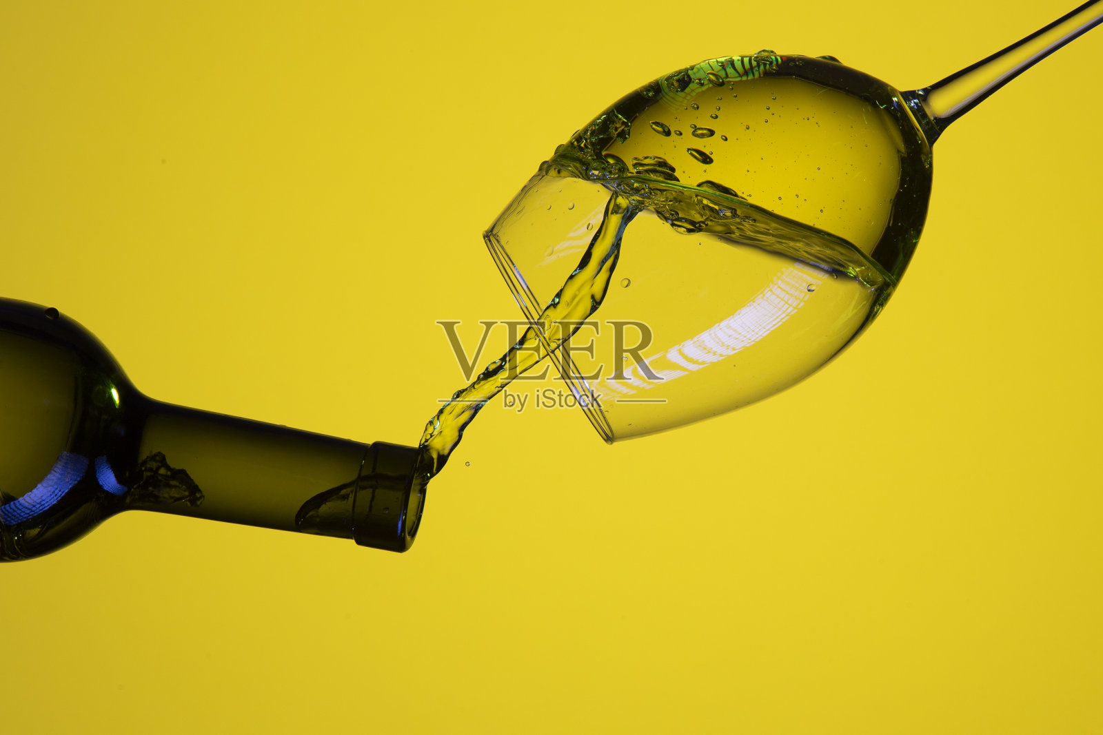 从瓶子中倒出彩色液体和水滴到酒杯中。在黄色背景上孤立。倒像照片摄影图片