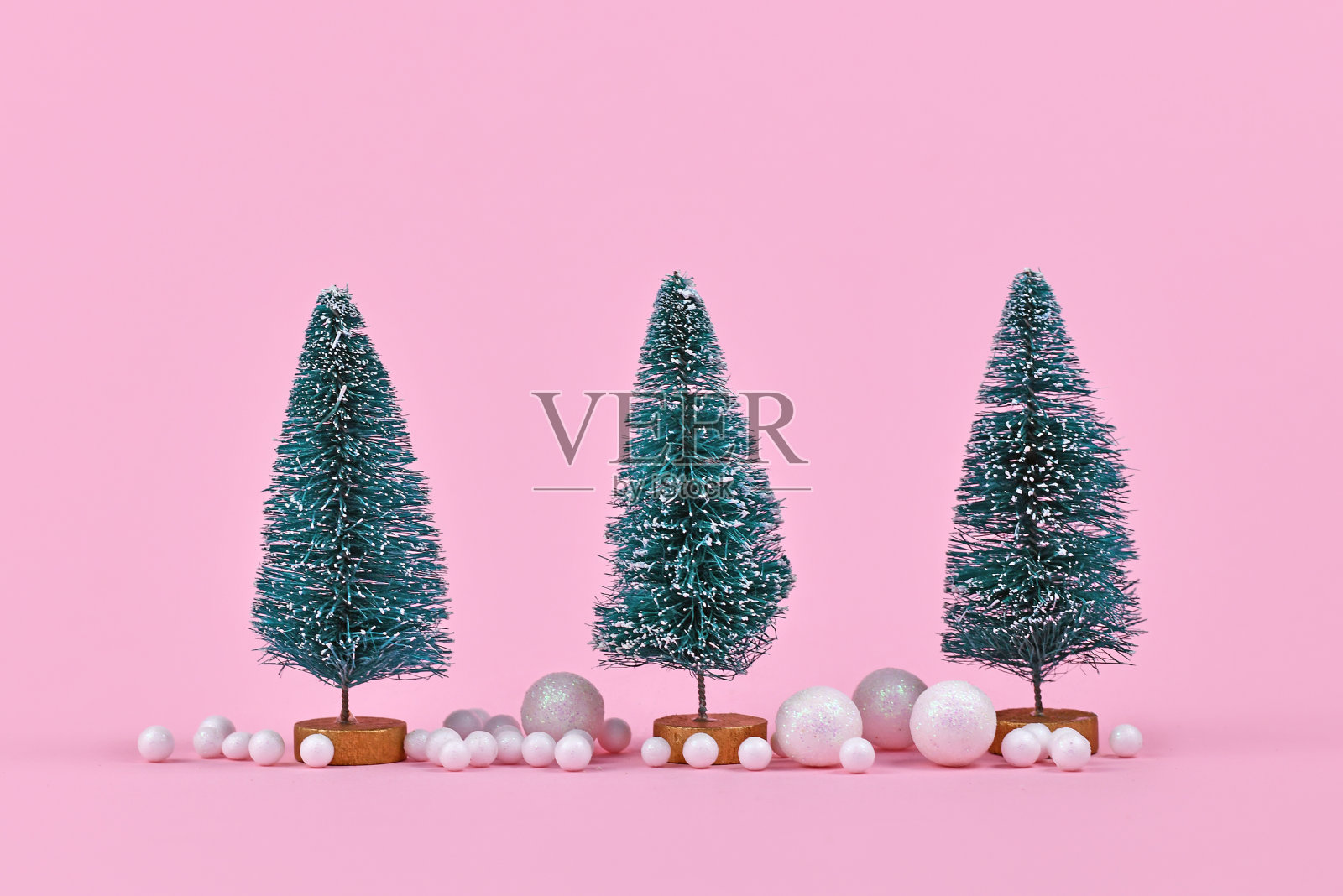 三棵小圣诞树被粉红色背景上的白色雪球装饰环绕着照片摄影图片