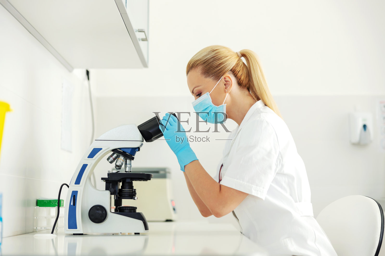 漂亮的实验室助理戴着橡胶手套和面罩坐在实验室里看槽状显微镜。照片摄影图片