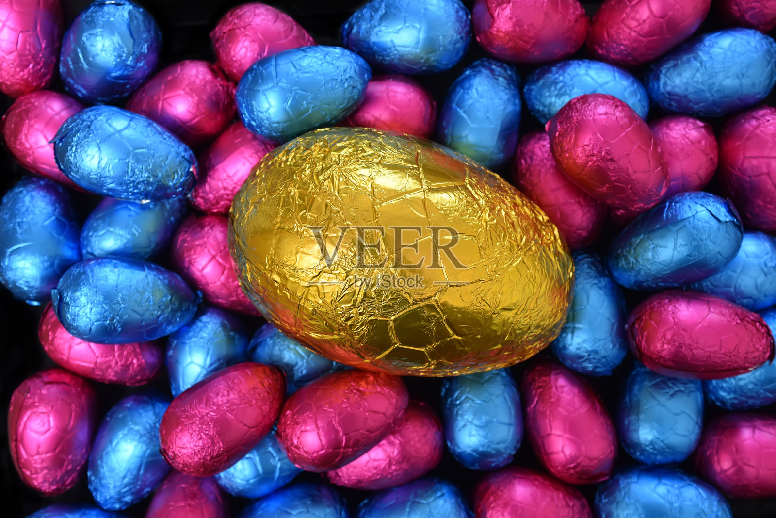 一堆或一组五颜六色、大小不一的用锡箔纸包裹的巧克力复活节彩蛋，其中有粉红色的、蓝色的，中间放一个大大的金色彩蛋。照片摄影图片