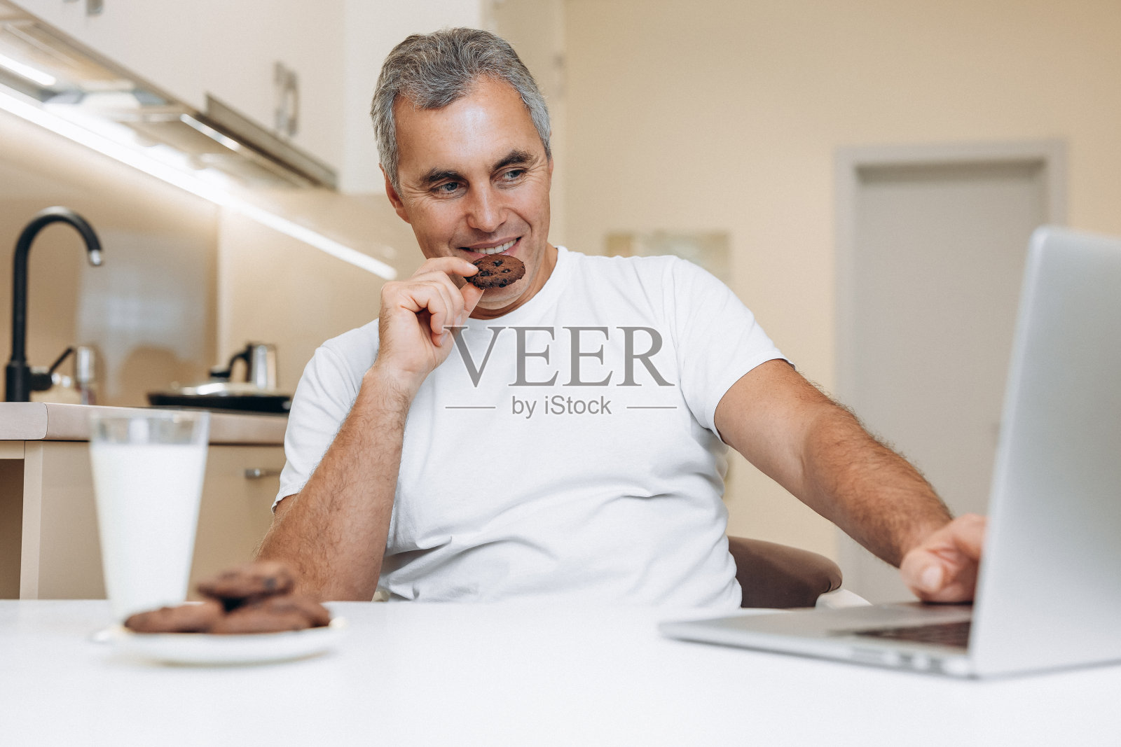 一个成熟的男人坐在厨房里，用着他的现代灰色笔记本电脑，看着搞笑的连续剧或视频，一边笑一边吃着美味的饼干。白色的桌子上放着一杯新鲜的牛奶和饼干。照片摄影图片