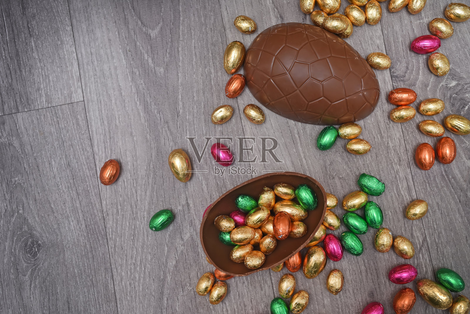 一堆或一组多颜色和不同大小的彩色锡箔包装的巧克力复活节蛋在粉红色，红色，金色和绿色。一个大的半块棕色牛奶巧克力鸡蛋，里面有迷你的鸡蛋，在灰色的木头背景上。照片摄影图片
