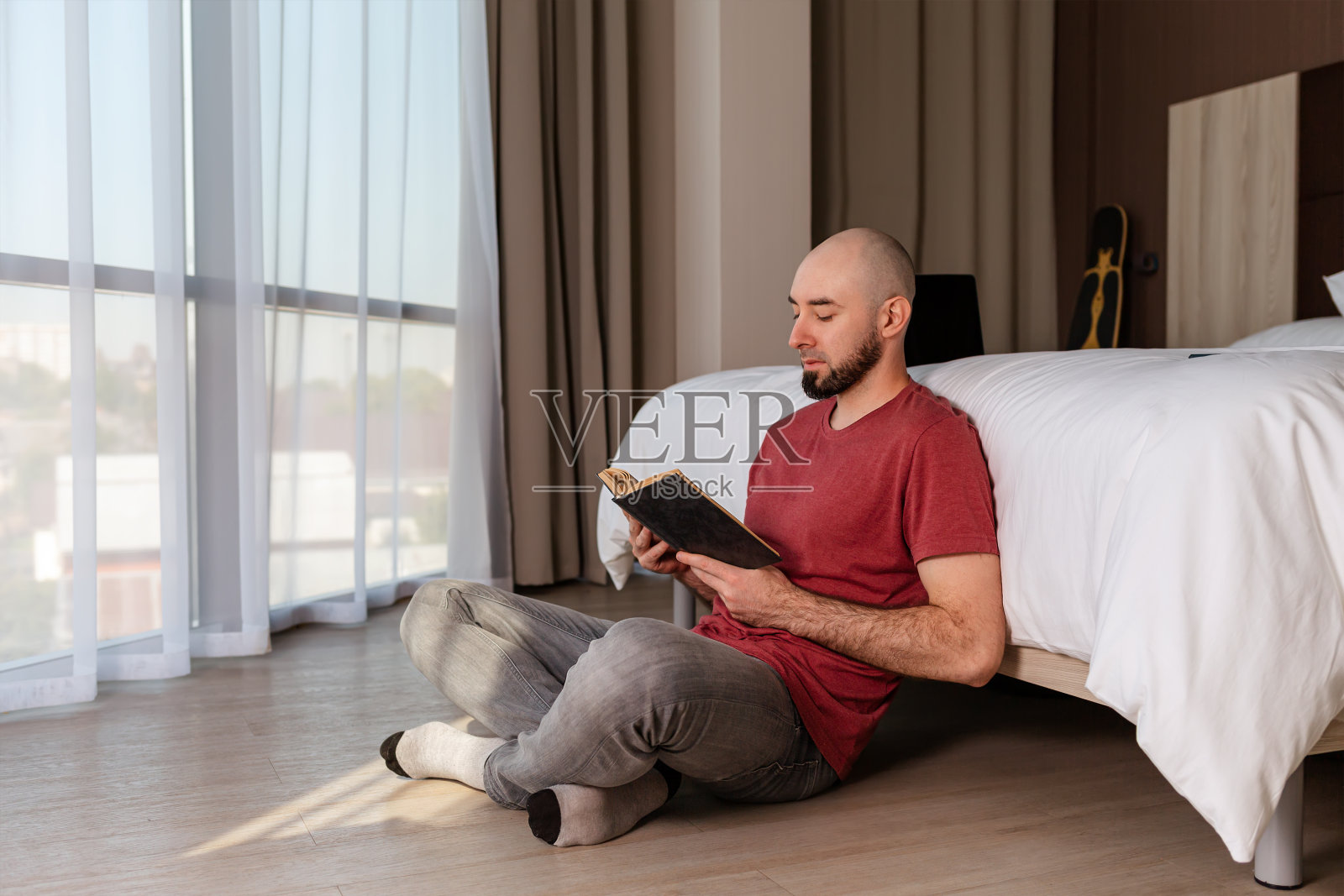 一个留着胡子的秃顶年轻人盘腿坐在他家的地板上看书。室内。教育与阅读的概念照片摄影图片
