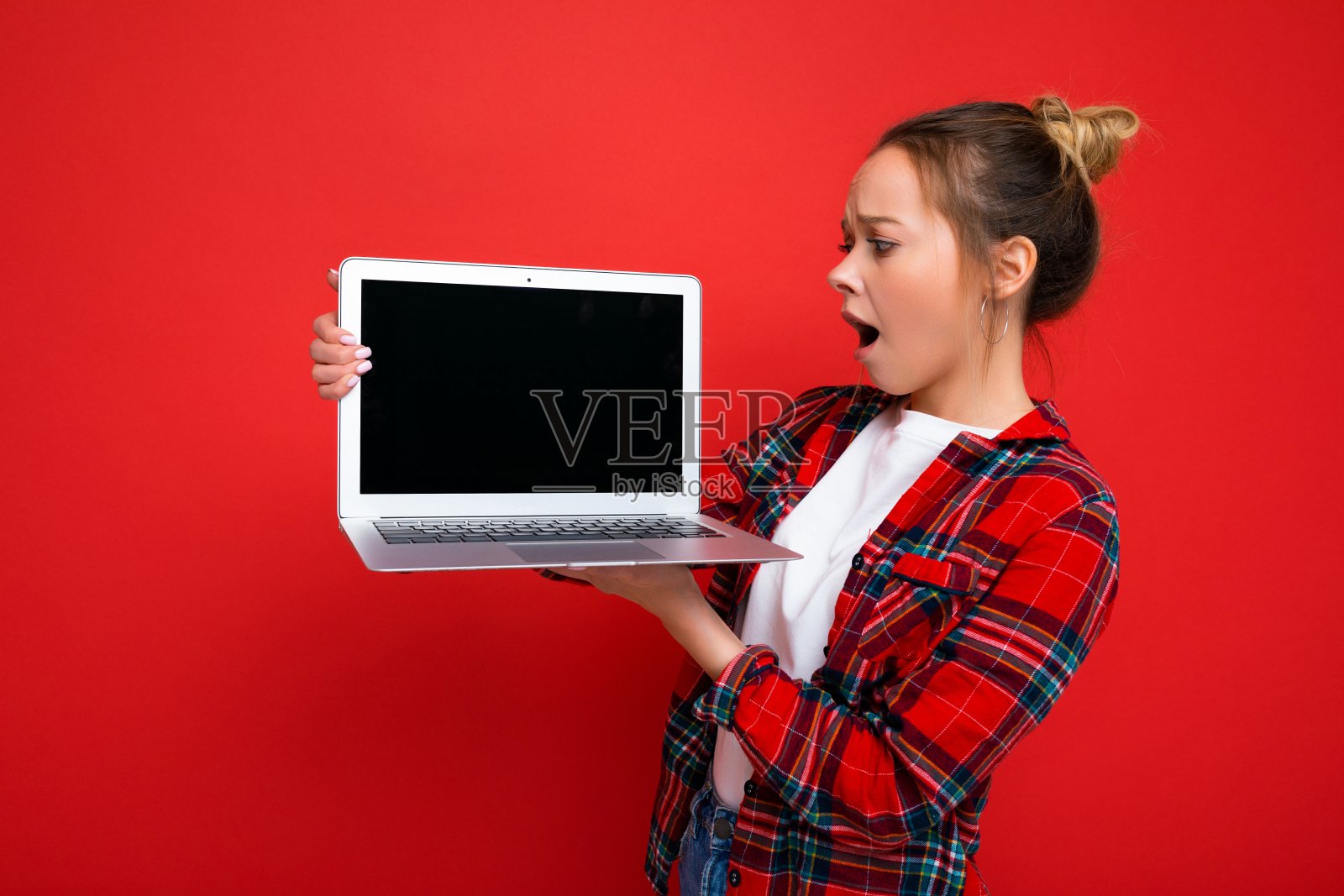 美丽震惊的年轻女子拿着笔记本电脑穿着红色衬衫看着显示器孤立在红色背景照片摄影图片