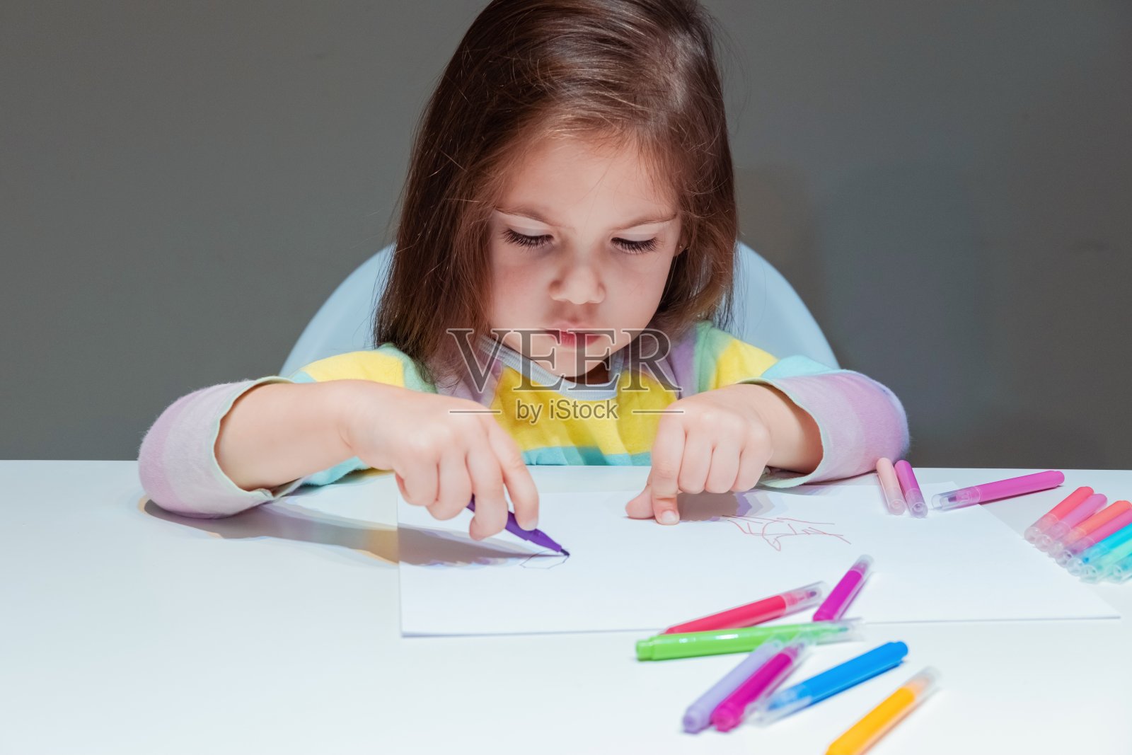 可爱的小女孩坐在桌子上，用毡尖笔在一张白纸上画画。灰色的背景。照片摄影图片
