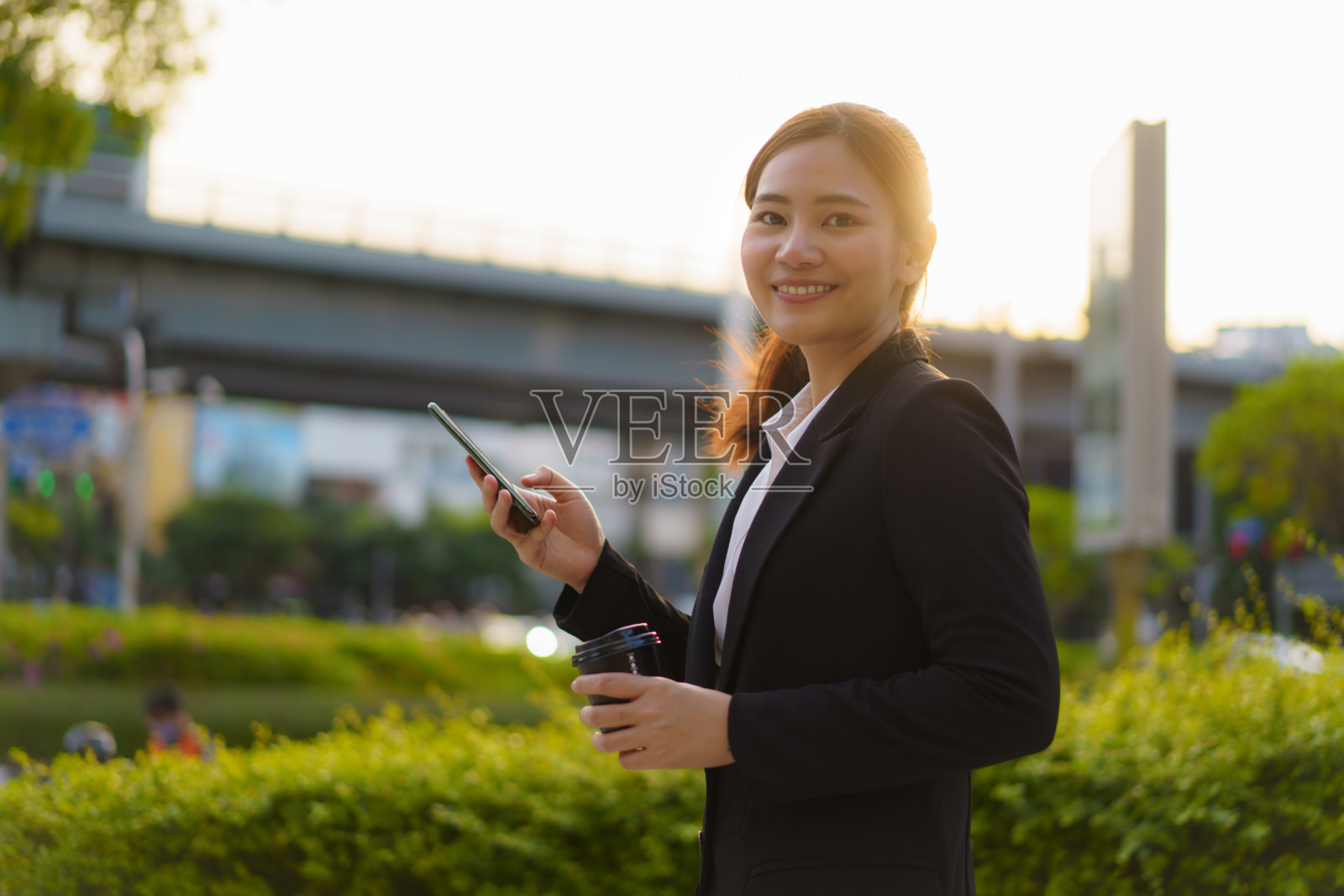 在泰国曼谷，一名亚洲行政工作女性拿着咖啡杯，在街道上使用手机，背景是办公大楼。照片摄影图片