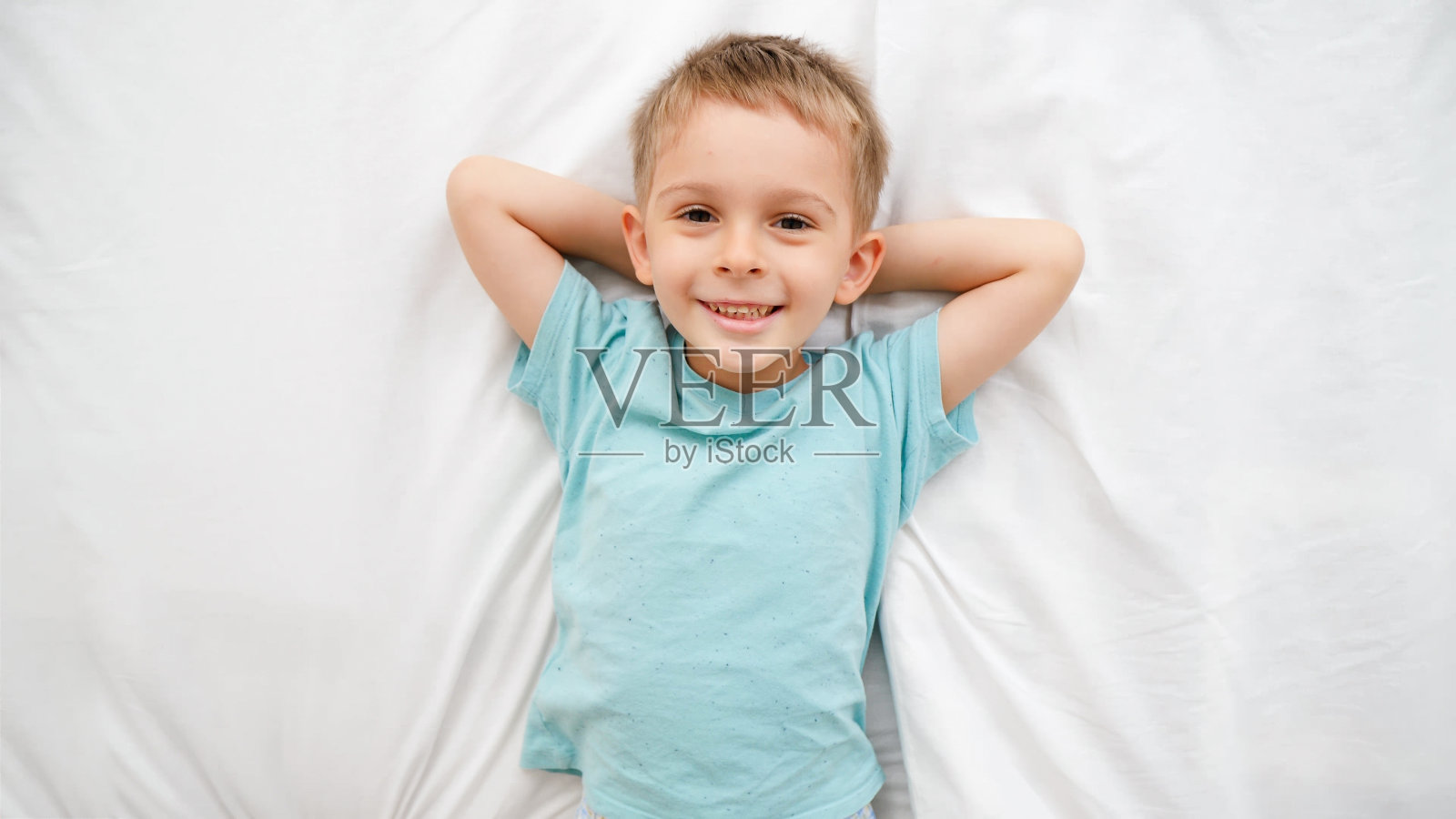 俯视图肖像的快乐微笑的蹒跚学步的男孩躺在床上，在镜头里笑。孩子们在家里玩得开心的概念照片摄影图片