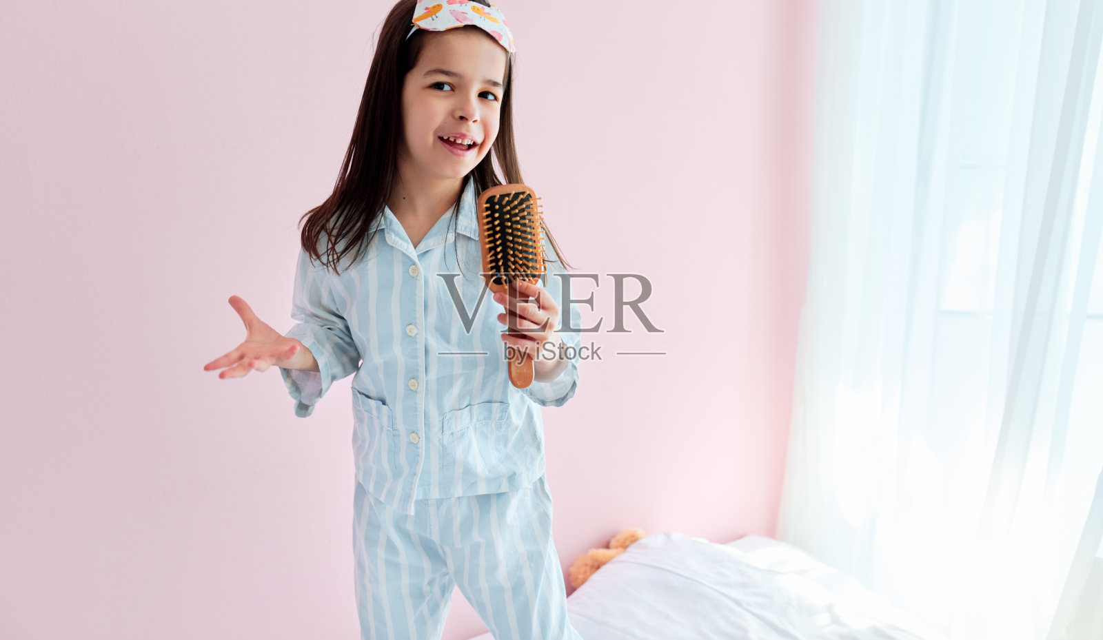 可爱的小女孩穿着睡衣在床上拿着梳子像麦克风唱歌模仿自己一个真正的歌手早上。照片摄影图片