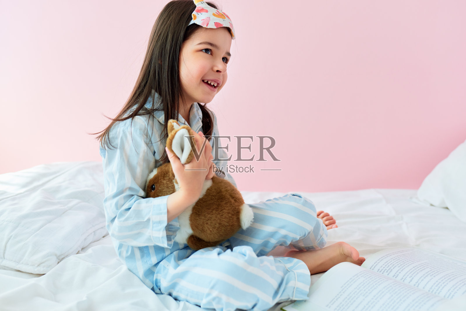 一个顽皮的小女孩穿着睡衣在床上玩玩具在现代舒适的阳光卧室的侧视图。快乐的孩子休息在床上在早上在家里在她的生日。照片摄影图片