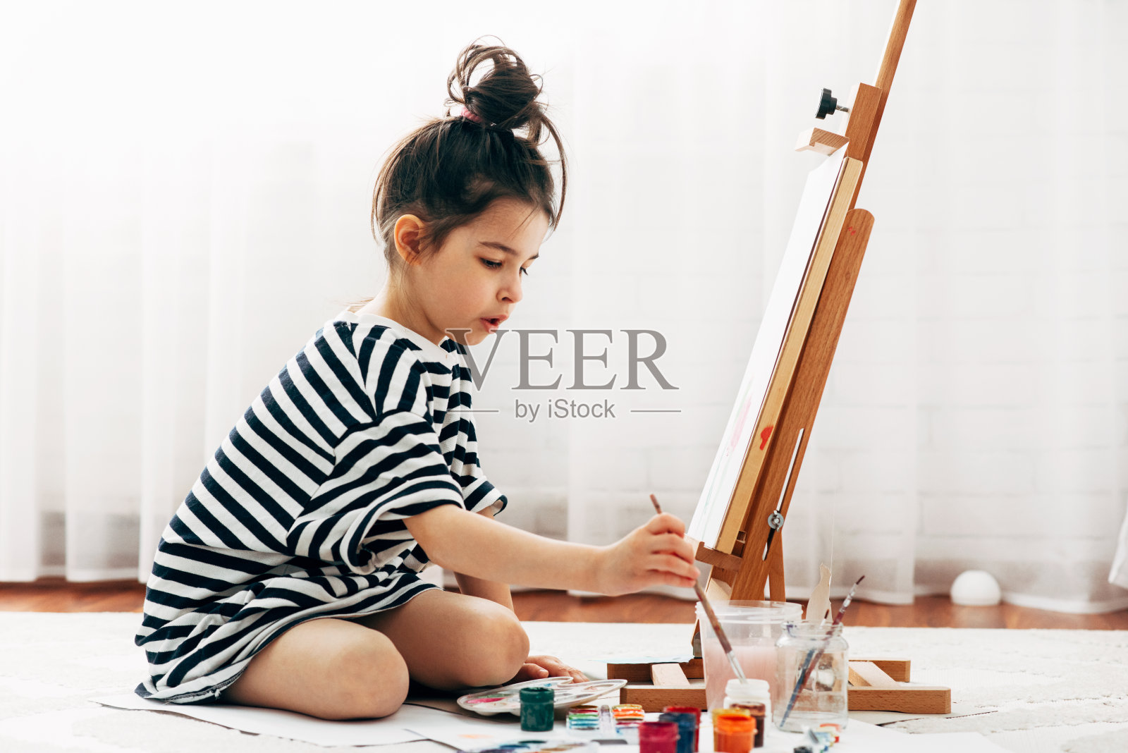 一个学龄前的小女孩在家里的画架上用画笔在纸上画的水平的全长图像。一个可爱的孩子在禁闭期间坐在地板上在她房间里画画。照片摄影图片
