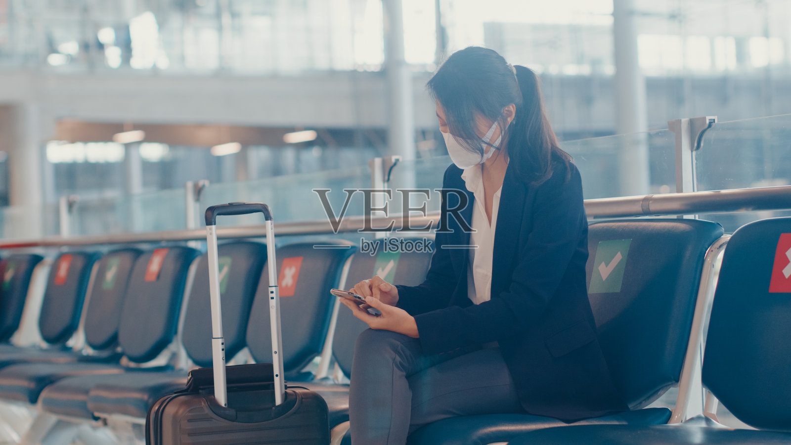 亚洲商务女旅行者穿着西装，拿着行李箱坐在机场的长椅上用智能手机聊天，等待航班。照片摄影图片