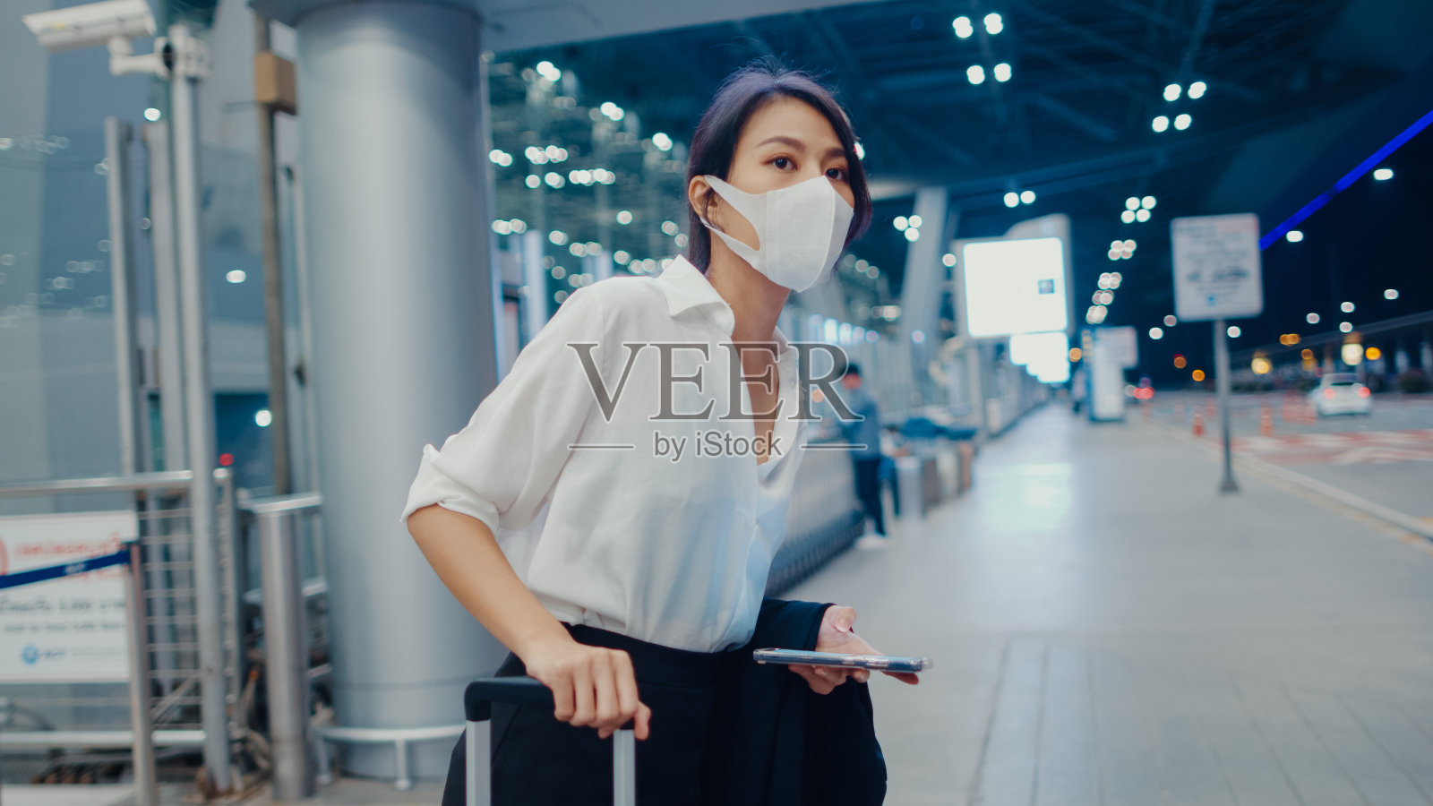 亚洲商务小姐到达目的地戴口罩站在外面看智能手机在国内机场候机楼等候汽车。照片摄影图片