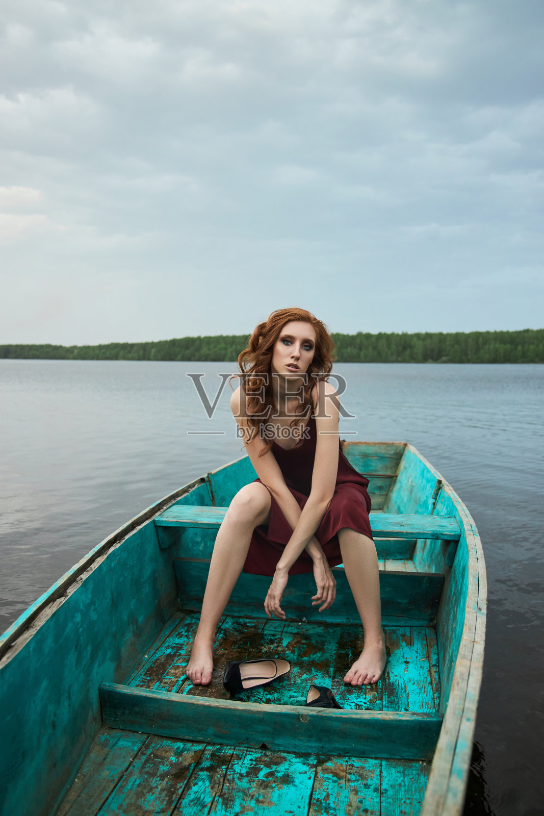 漂亮时尚的红发女人坐在船上。美丽浪漫的肖像女孩在湖上的木船上穿着红色葡萄酒的裙子照片摄影图片