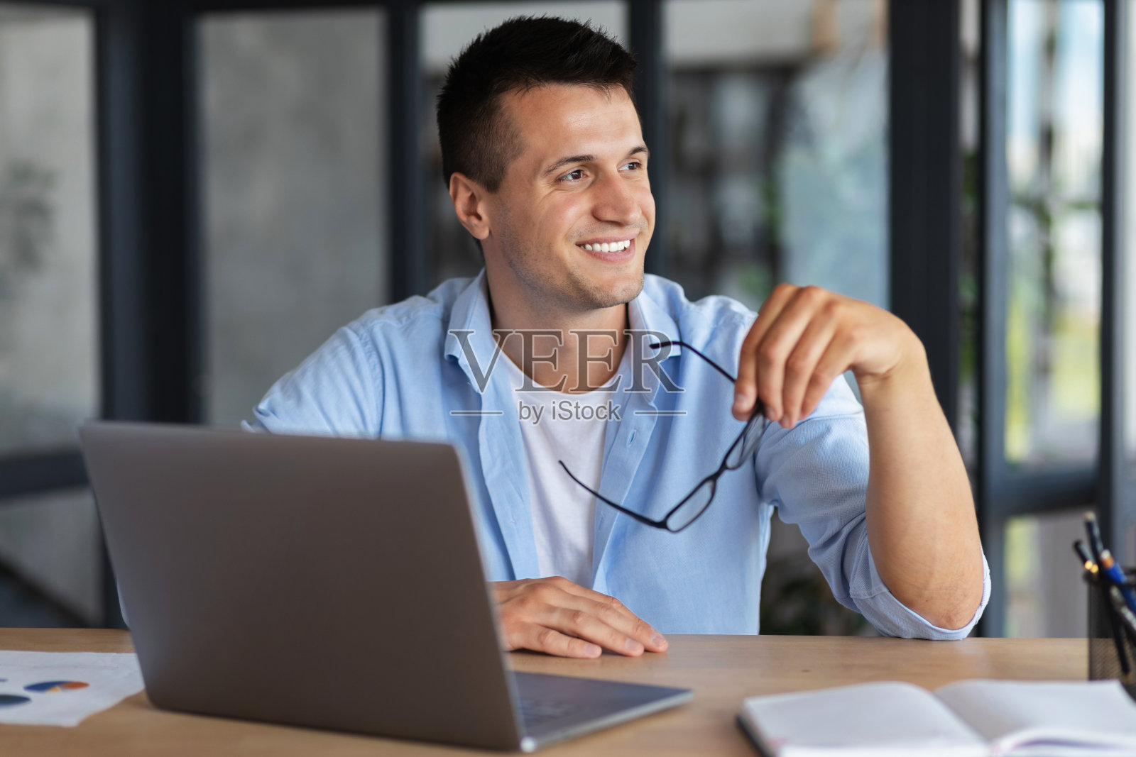快乐的年轻白人商人或学生，坐在桌旁的笔记本电脑看向一边和可爱的微笑，男性思考未来的计划照片摄影图片