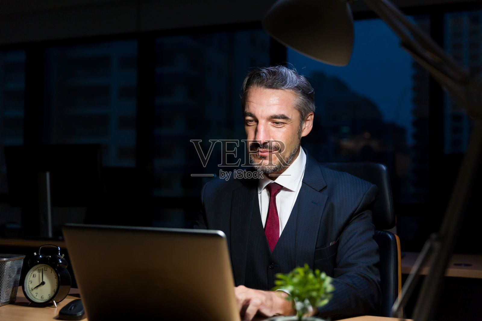 商人、办公室职员穿着正式的西装，晚上在黑暗的办公室里坐在办公桌前工作。商人在灯光下用笔记本电脑加班照片摄影图片