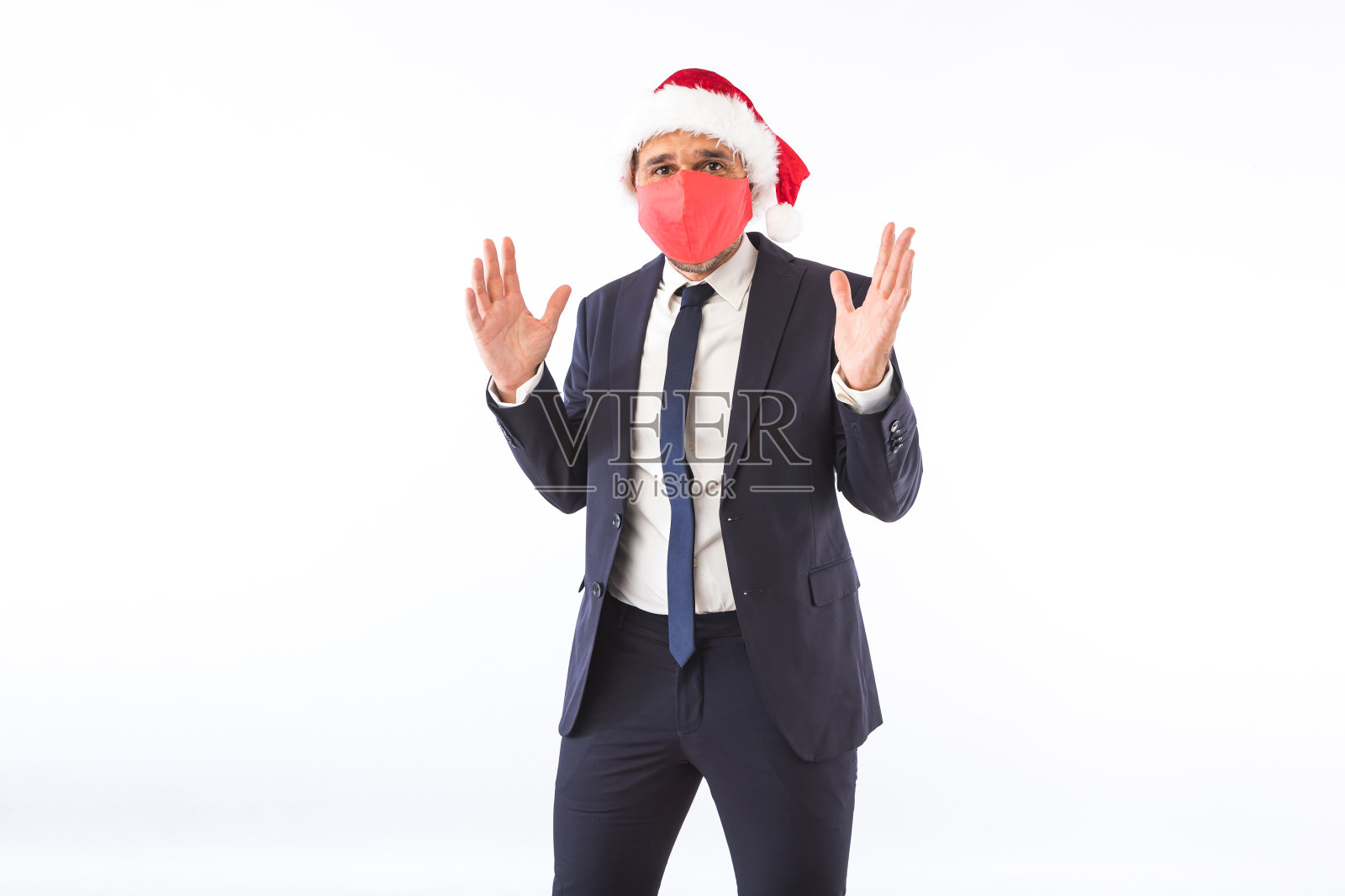 商人穿着西装，打着领带，圣诞老人戴着圣诞帽，戴着红色面具，在白色的背景上举起手来玩。圣诞节庆祝活动的概念。照片摄影图片
