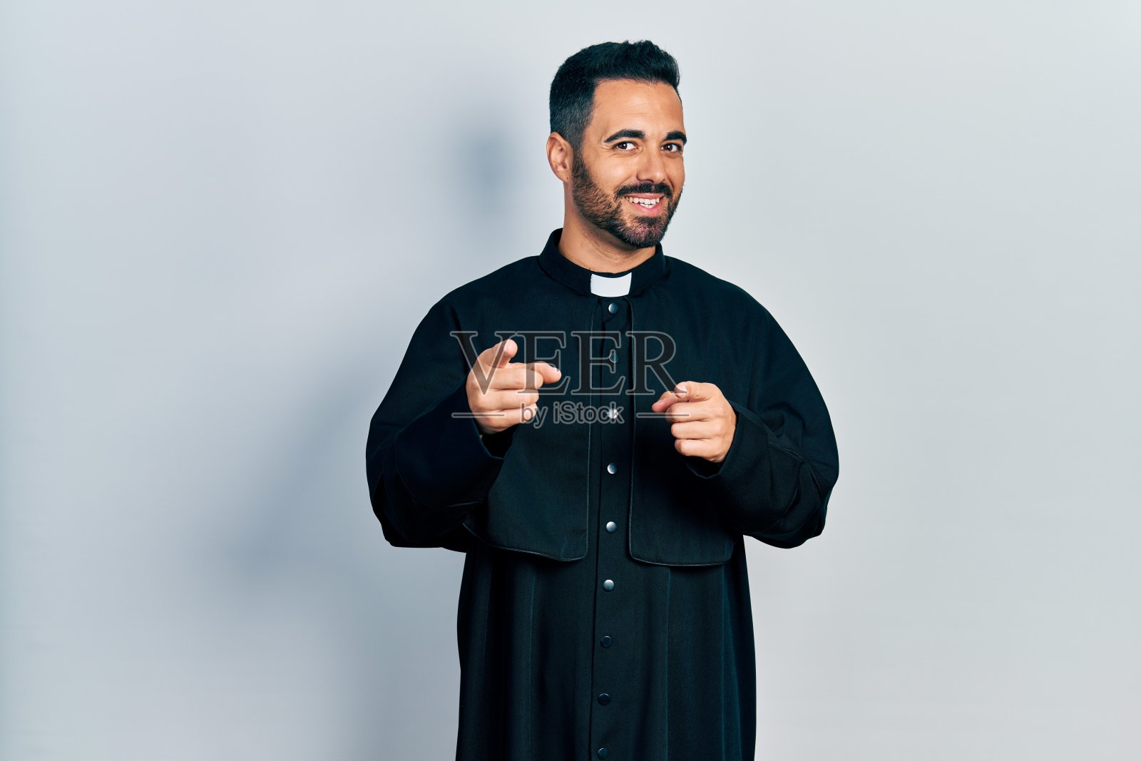 英俊的西班牙裔男子，留着大胡子，身穿天主教神父长袍，笑容可掬地指着镜头。良好的能量和氛围。照片摄影图片
