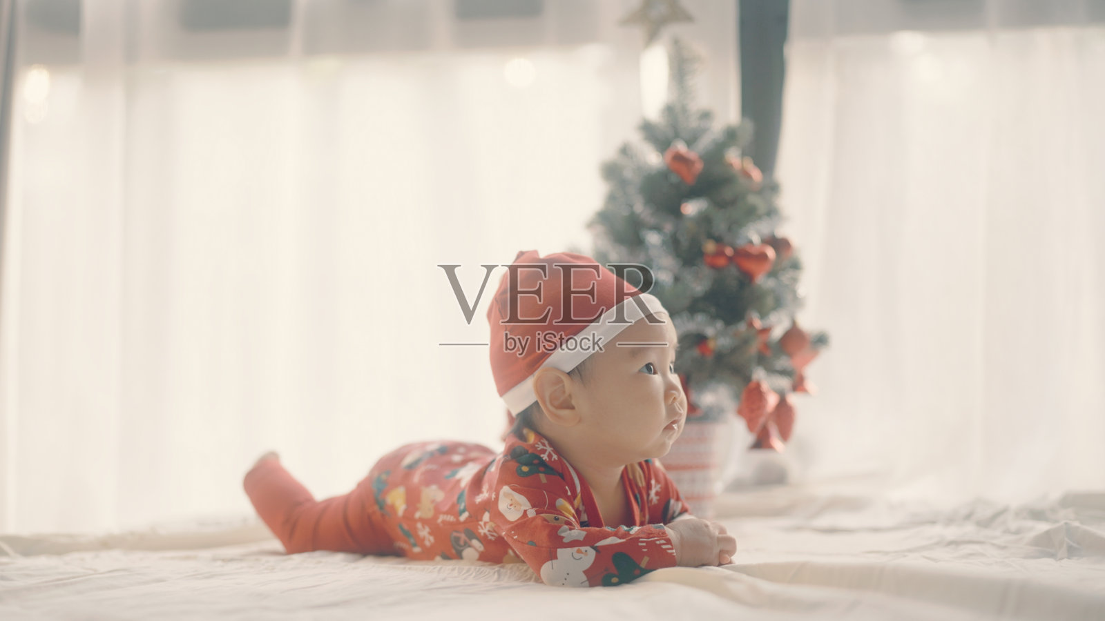 可爱的小婴儿戴着圣诞帽在圣诞树旁。照片摄影图片