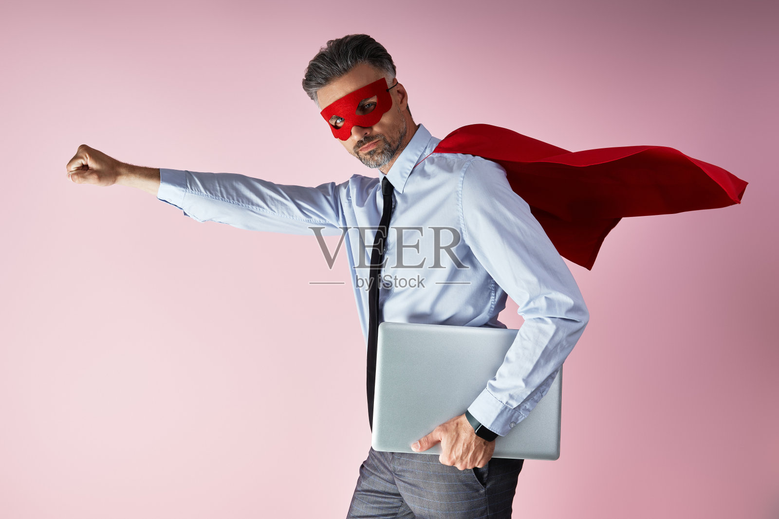 自信的男人穿着衬衫和领带，穿着超级英雄斗篷，在粉色背景下拿着笔记本电脑照片摄影图片